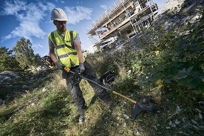 Ein Mann in Arbeitskleidung der eine Dewalt DCM571N 54 Volt Akku-Motorsense auf einer mit Unkraut bewachsenen Wiese benutzt